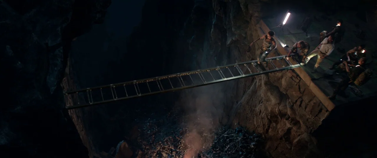 Tomb Raider (2018) movie still: wide — A group of people stands on a narrow stone ledge overlooking a deep rocky chasm. A woman crawls across a…