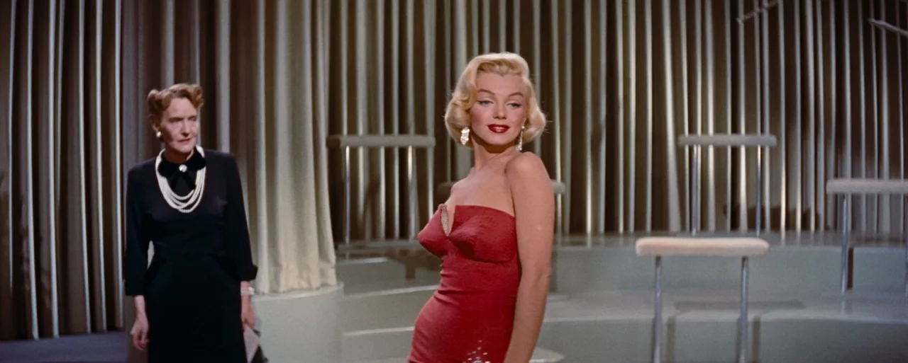 How to Marry a Millionaire movie still: medium — A theatrical stage features a metallic vertical-slat background and curved tiered platforms. A…