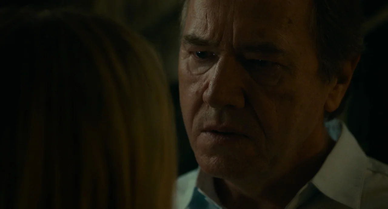 Last Summer (2023) movie still: closeup — An older man stands in a dimly lit indoor space with low ambient light. He looks intently toward a person…
