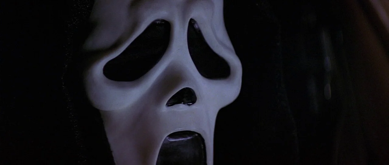 Scream 2 (1997) movie still: closeup — A dark, nondescript area provides the backdrop for a central close-up focus. The white mask with distorted…