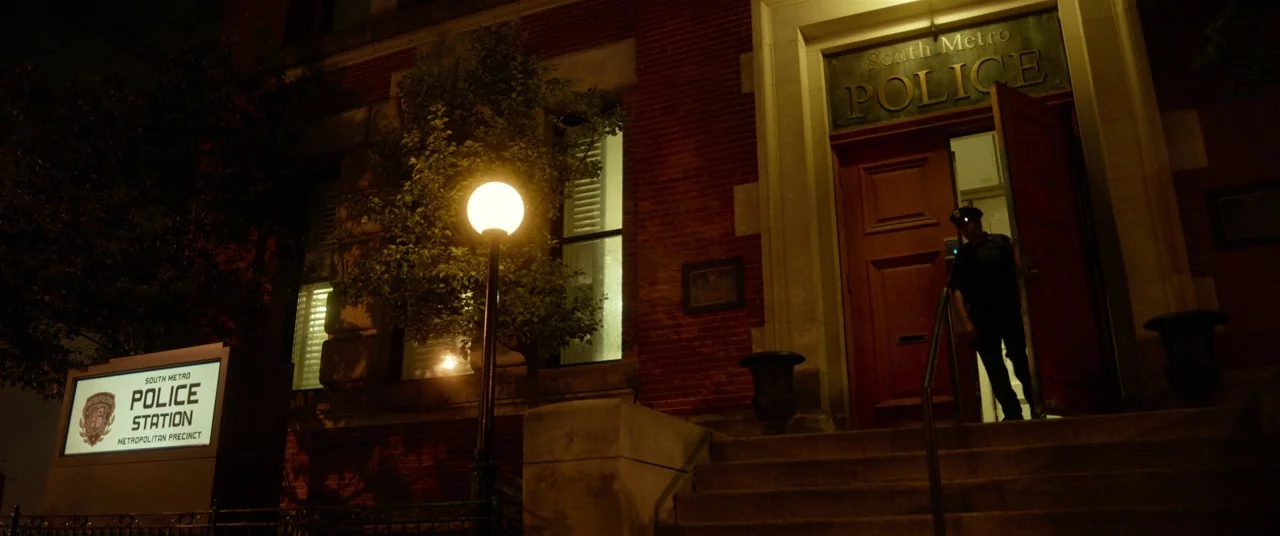 Spiral (2021) movie still: wide — A brick police station facade features a wide wooden door under a lit sign. A uniformed police officer stands in…