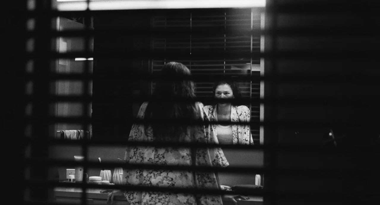 Malcolm & Marie (2021) movie still: medium — A woman stands before a vanity mirror in a dimly lit, tiled environment. The camera view is obscured by…