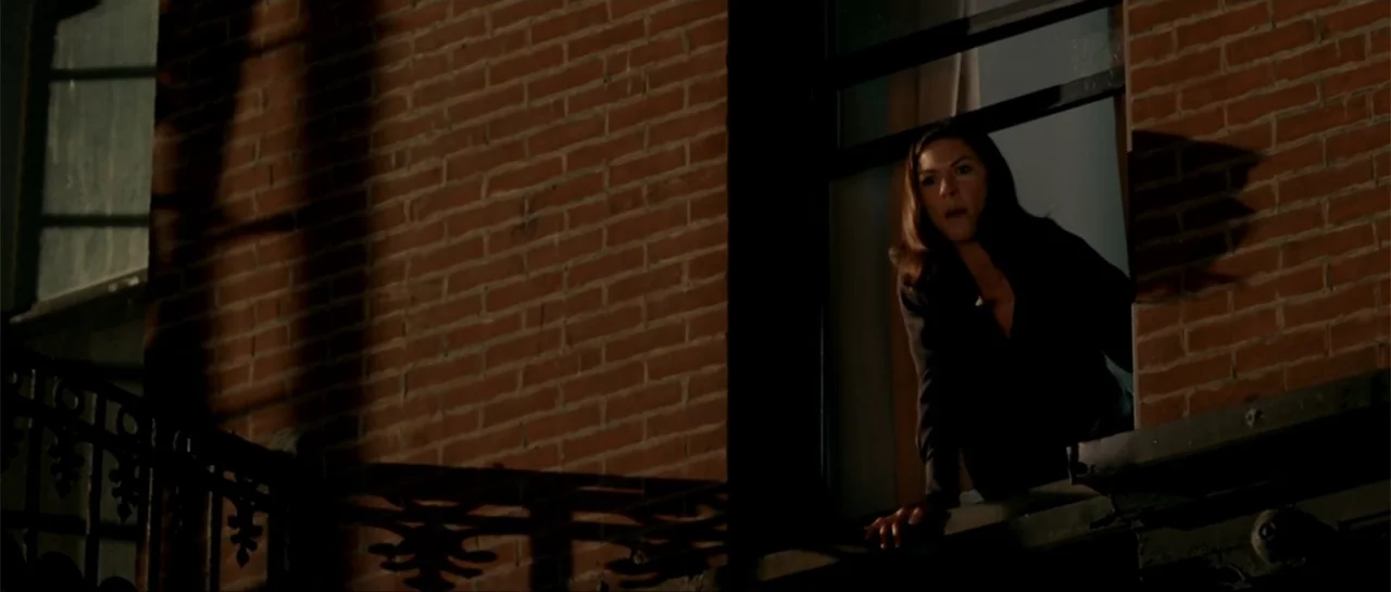 The Rebound (2009) movie still: medium — A dark brick building exterior at night features a woman looking out from an open window. The woman reaches…