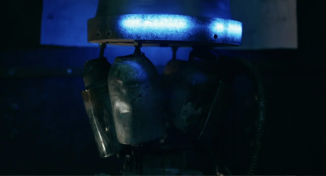 The Lift (2021) movie still: closeup — A close-up view of a metallic machine component with blue light emitting from above. The dark background…