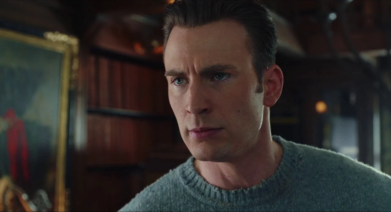 Knives Out (2019) movie still: closeup — A dimly lit room features bookshelves filled with books and a framed painting. A man wears a thick grey…