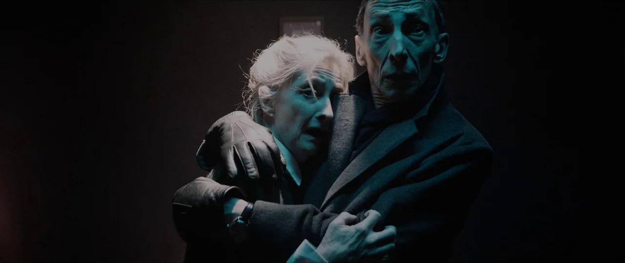 Anything For Jackson (2020) movie still: medium — An elderly couple in dark clothing is shown in a dimly lit, undefined interior space. The woman…