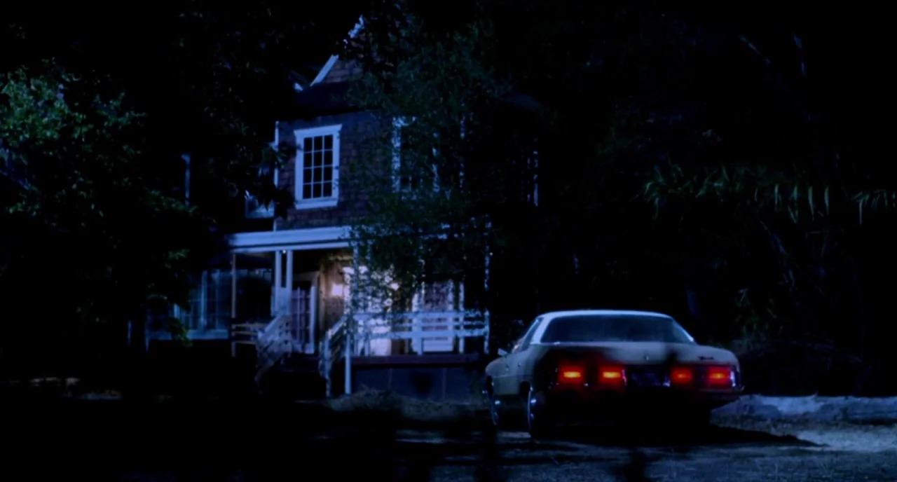 The Love Witch (2016) movie still: establishing — A two-story house with a wooden porch stands surrounded by dark trees at night. A vintage sedan is…
