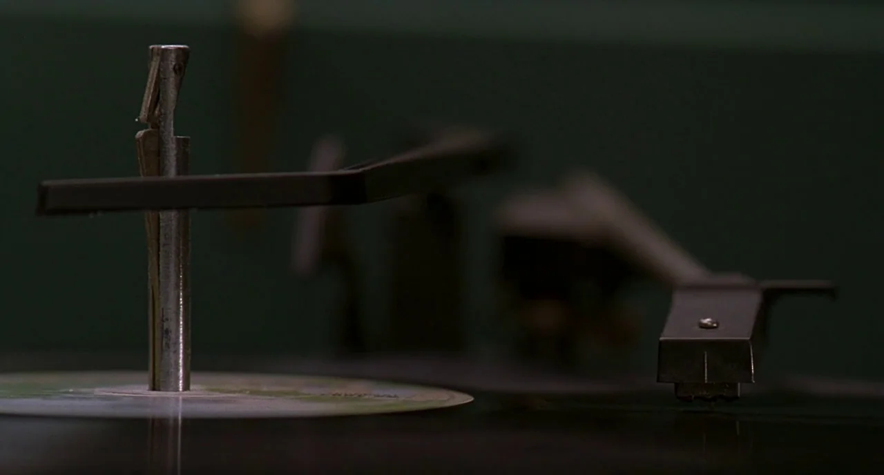 The Ice Storm (1997) movie still: closeup — A dark, close-up view of a vintage record player mechanism in a dim room. The metal central spindle and…