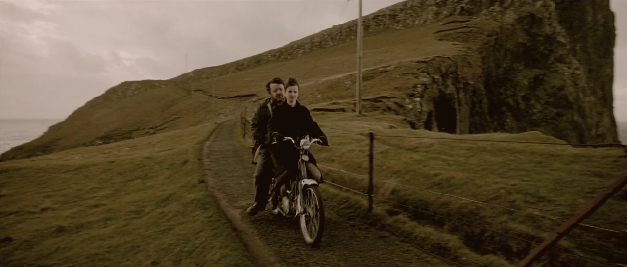 Breaking the Waves (1996) movie still: fullbody — A man and woman ride a motorbike on a narrow grass-lined gravel path. Rolling hills and steep…