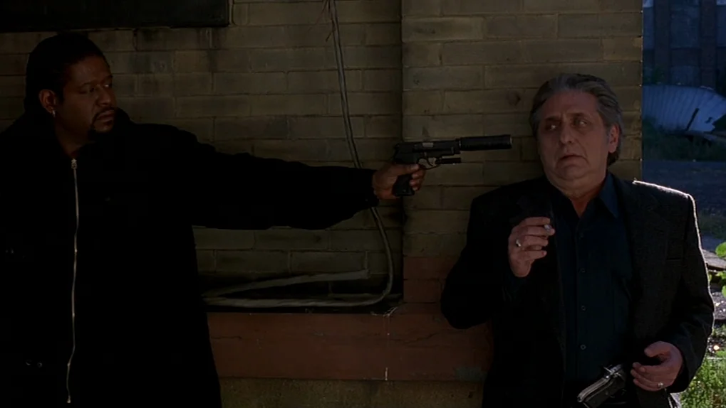Ghost Dog: The Way of the Samurai (1999) movie still: medium — A man in a black jacket stands beside a weathered yellow brick building. He aims a…