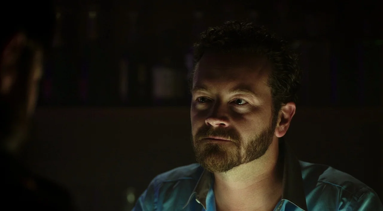 Urge (2016) movie still: closeup — A dimly lit bar environment with bottles visible as blurred shapes in the background. A man with a dark beard…