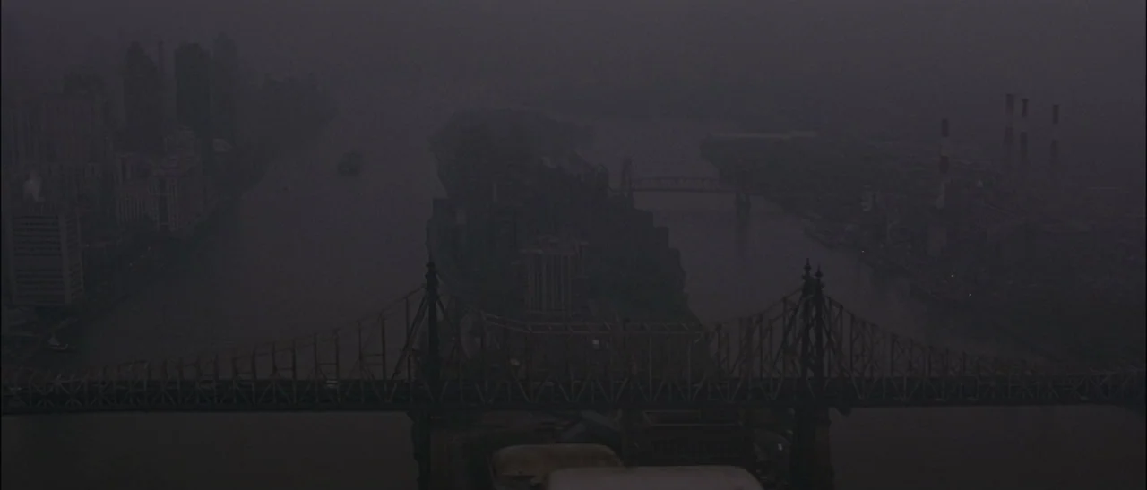 Dark Water (2005) movie still: establishing — A dark, foggy urban landscape features a large cantilever bridge spanning across a river. Industrial…
