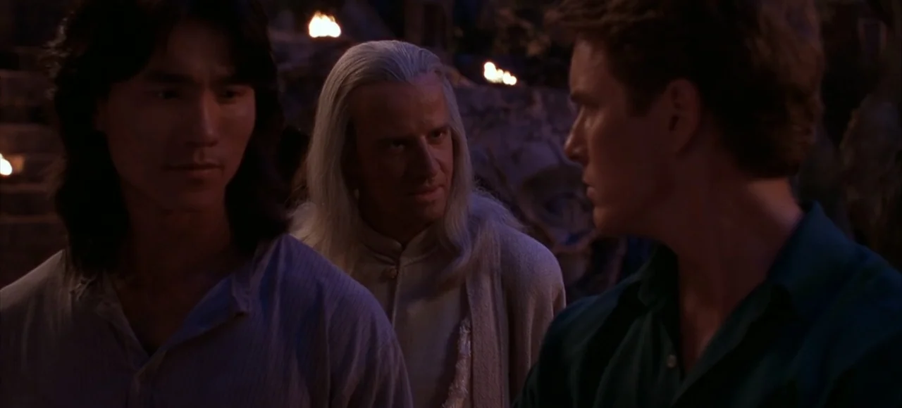 Mortal Kombat (2021) movie still: medium — An outdoor area with dark stone walls and flickering torches in the background. Three men stand together…