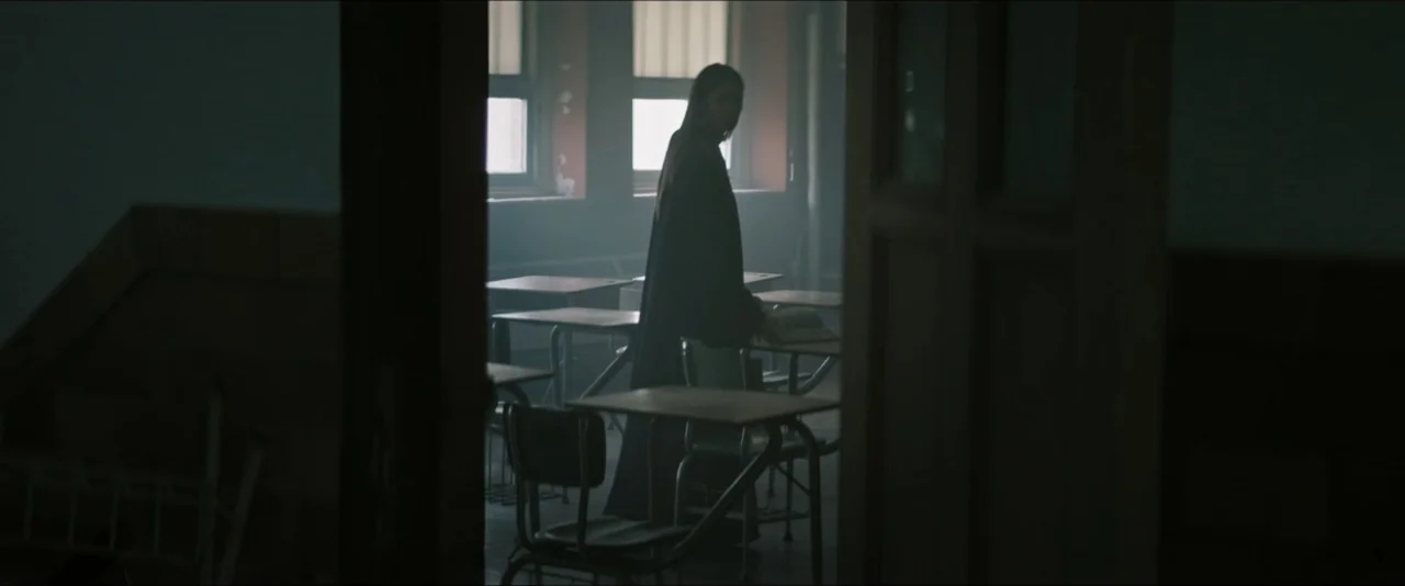 It Feeds (2025) movie still: fullbody — A dimly lit classroom features rows of empty student desks under grey window light. A woman in a dark coat…