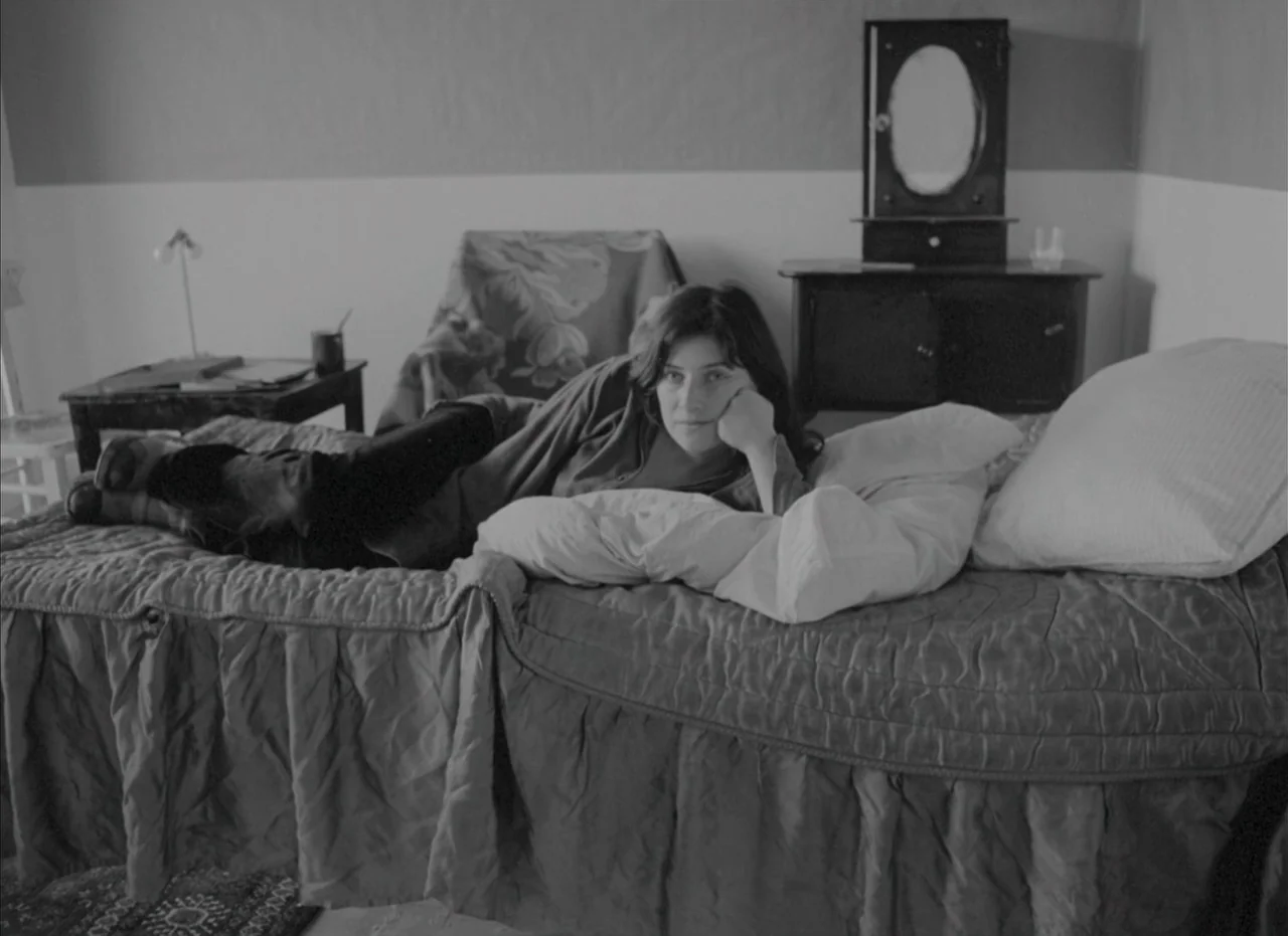 Je Tu Il Elle (1974) movie still: medium — A furnished bedroom features a bed with a quilted cover and a wall mirror. The woman rests on her side…