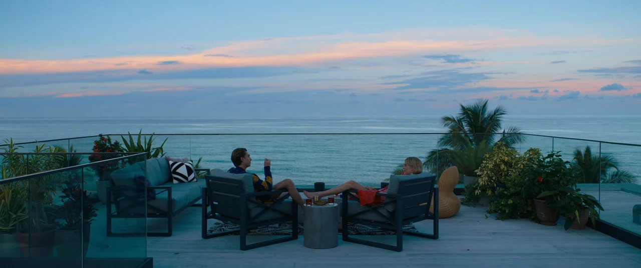 Do Revenge (2022) movie still: wide — A modern outdoor deck with grey lounge furniture overlooks a vast ocean at twilight. A man and a woman sit in…