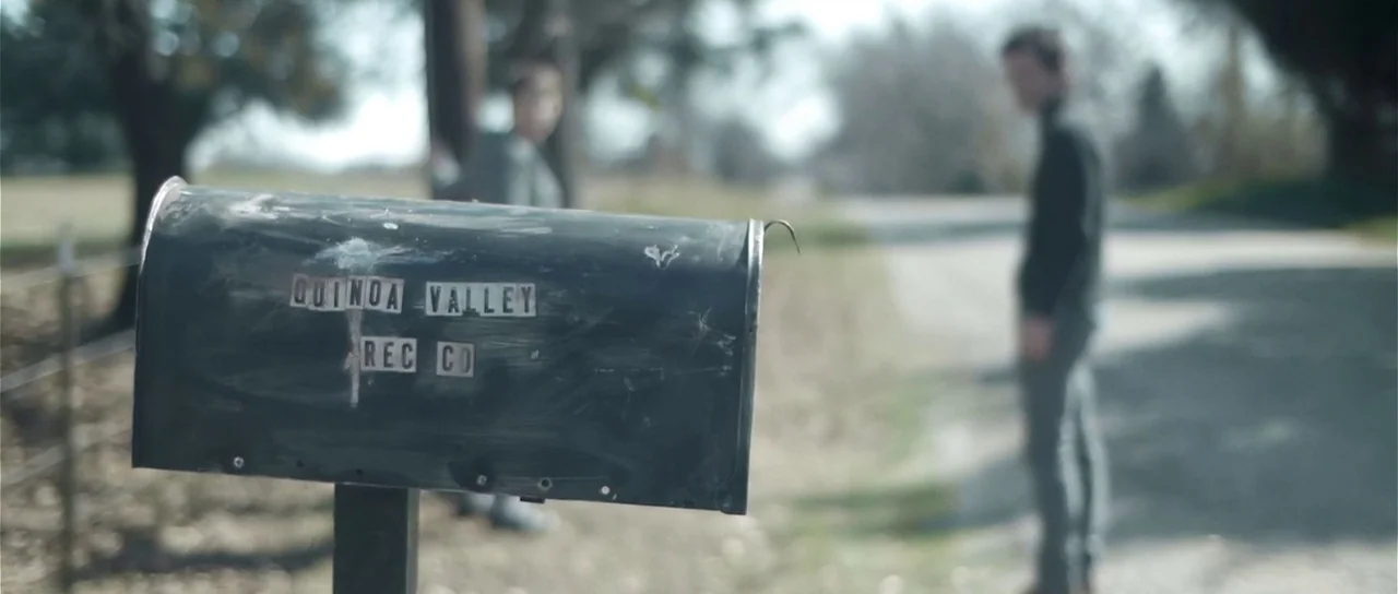 Upstream Color (2013) movie still: closeup — A rural road stretches into the distance with blurred figures standing near the roadside. In the…
