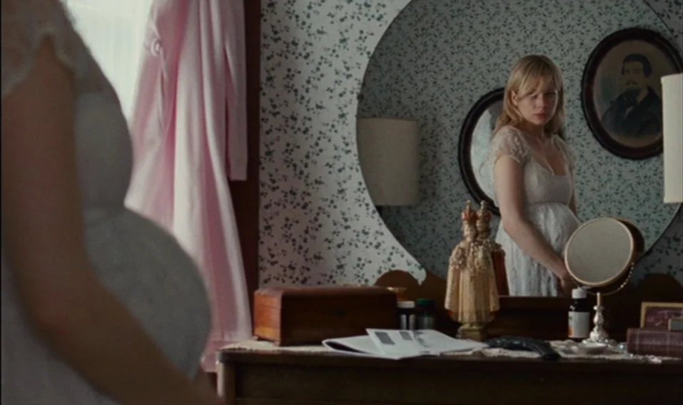 Blue Valentine (2010) movie still: medium — A woman stands before a vanity in a room with floral wallpaper and period furniture. She looks at her…