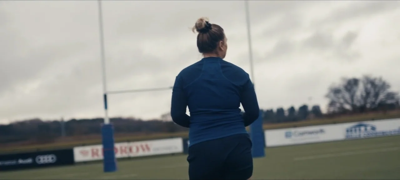 No Woman No Try (2022) movie still: medium — An outdoor sports field features white goalposts and perimeter advertising boards under an overcast…