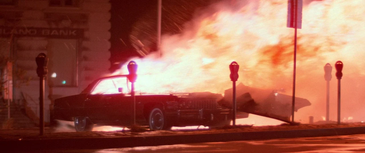 Vigilante (1982) movie still: wide — A dark city street features a burning vintage sedan parked by a bank building. Large flames erupt from the car…
