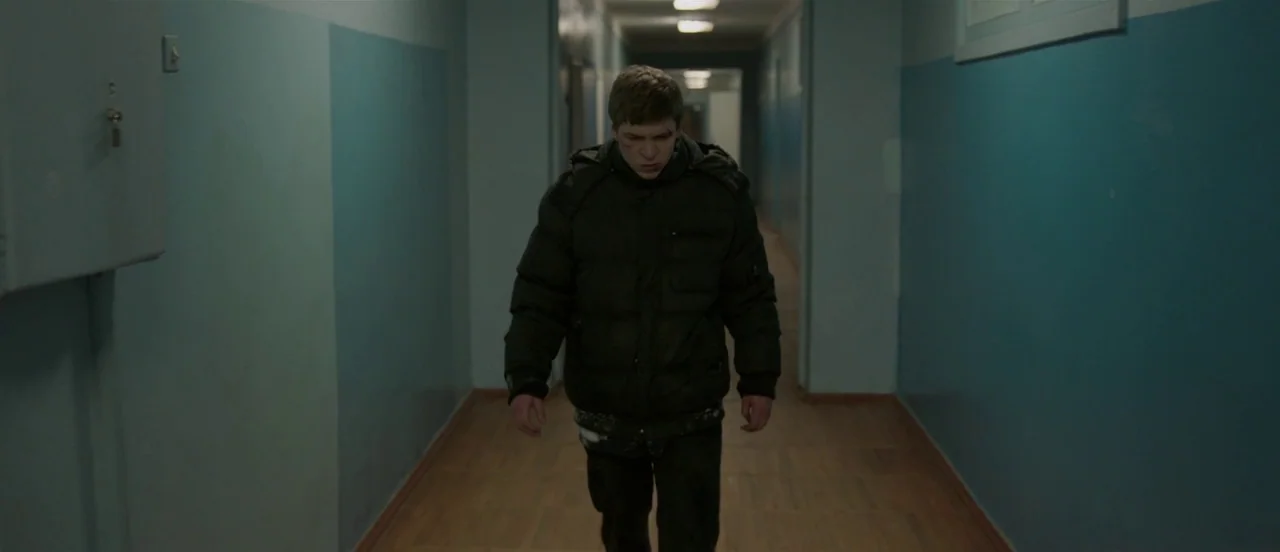 The Tribe (2014) movie still: medium — A long, dimly lit institutional passage with blue painted walls and wooden floorboards. A man in a dark…