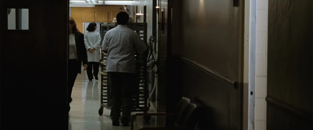 Conspiracy Theory (1997) movie still: medium — A linoleum-floored hospital corridor with beige walls and closed doors. An employee pushes a wheeled…