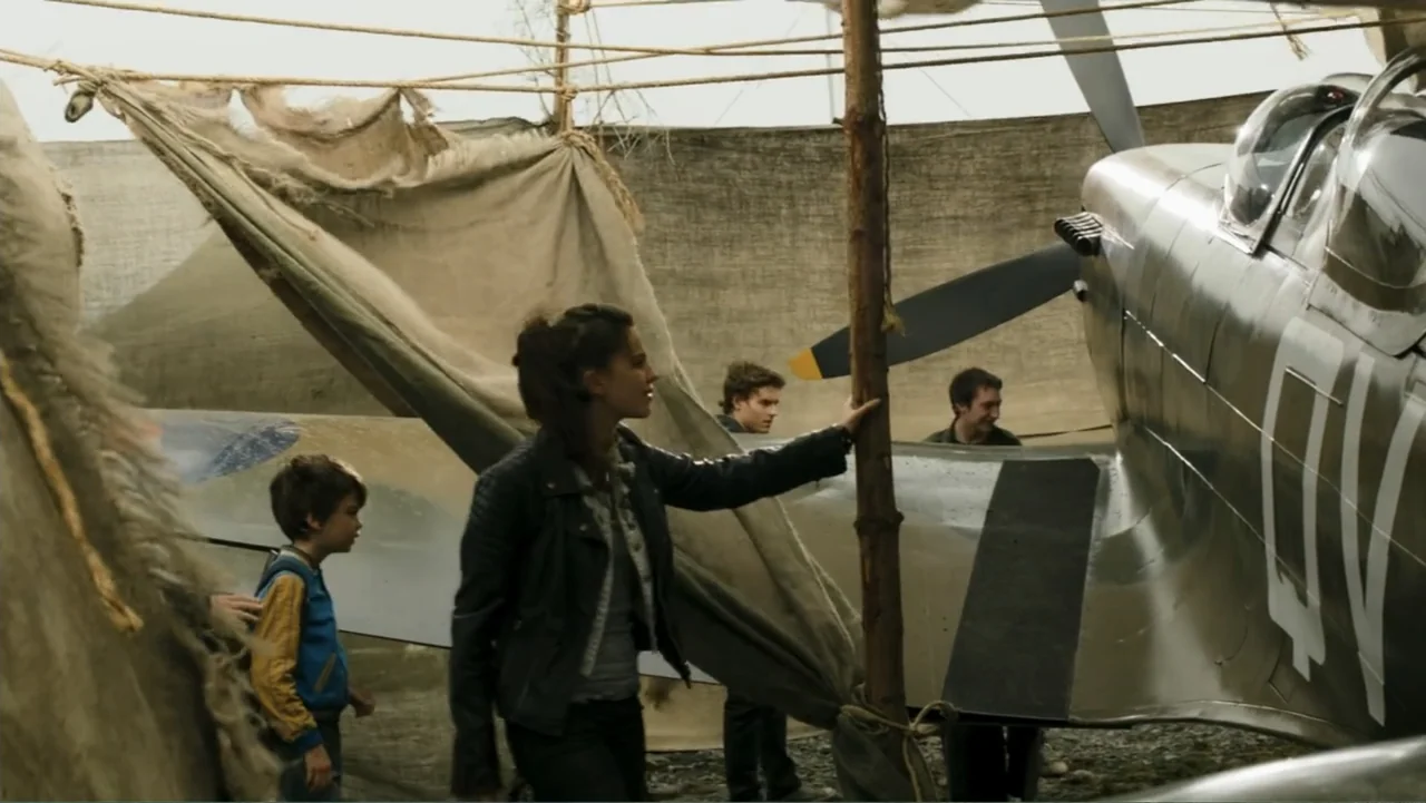 Robot Overlords (2015) movie still: medium — A makeshift hangar structure features a World War II fighter plane under burlap shade. A woman and a…