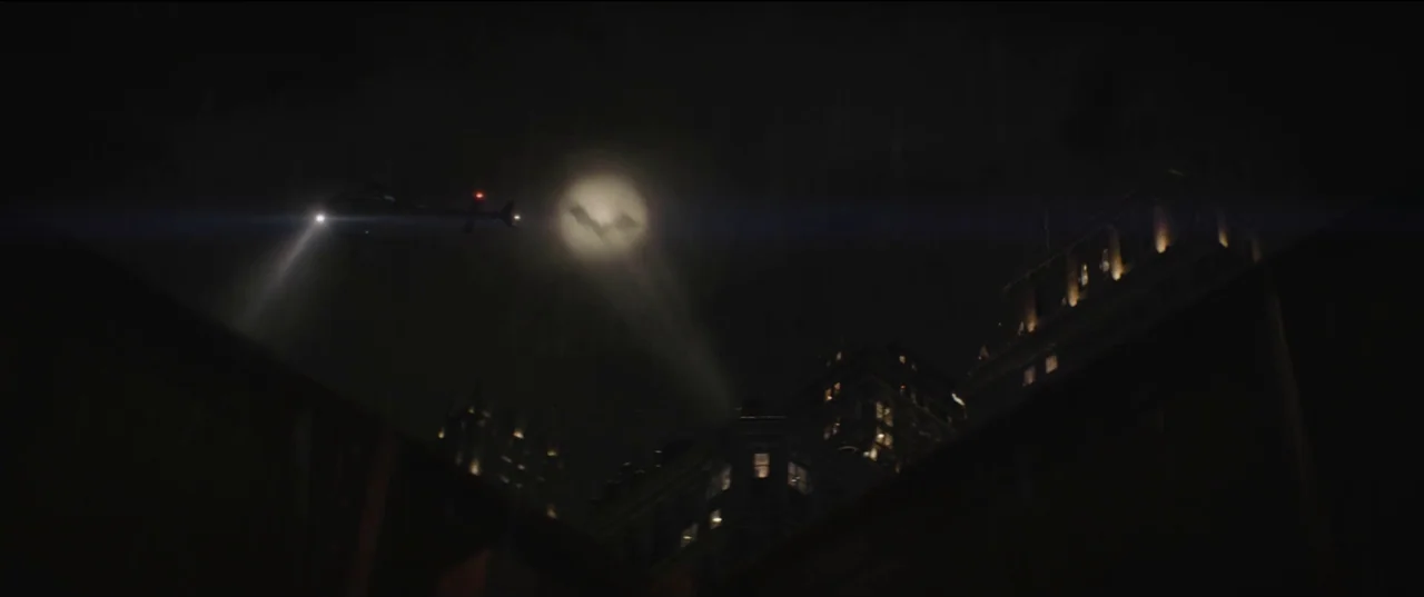 The Batman (2022) movie still: wide — A dark urban skyline at night features tall buildings illuminated by faint indoor lighting. A lone helicopter…