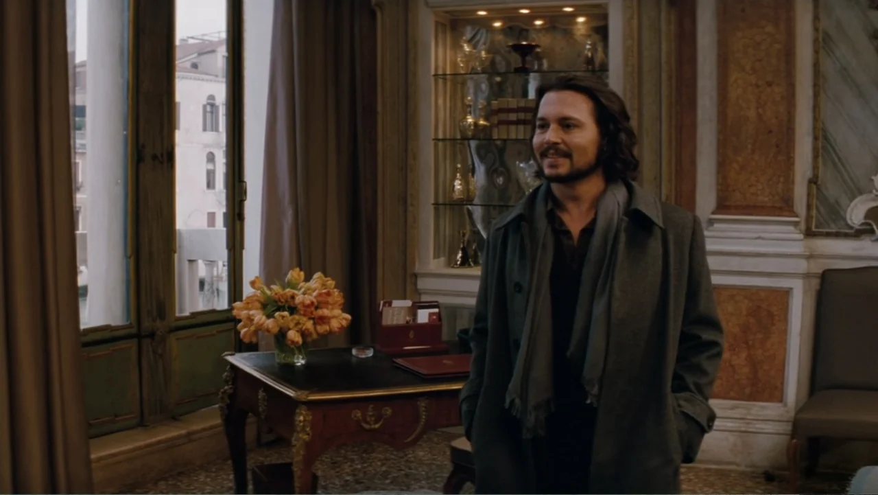 The Tourist (2010) movie still: medium — An opulent hotel room features wood paneling and a desk with fresh flowers. A man stands near the windowed…