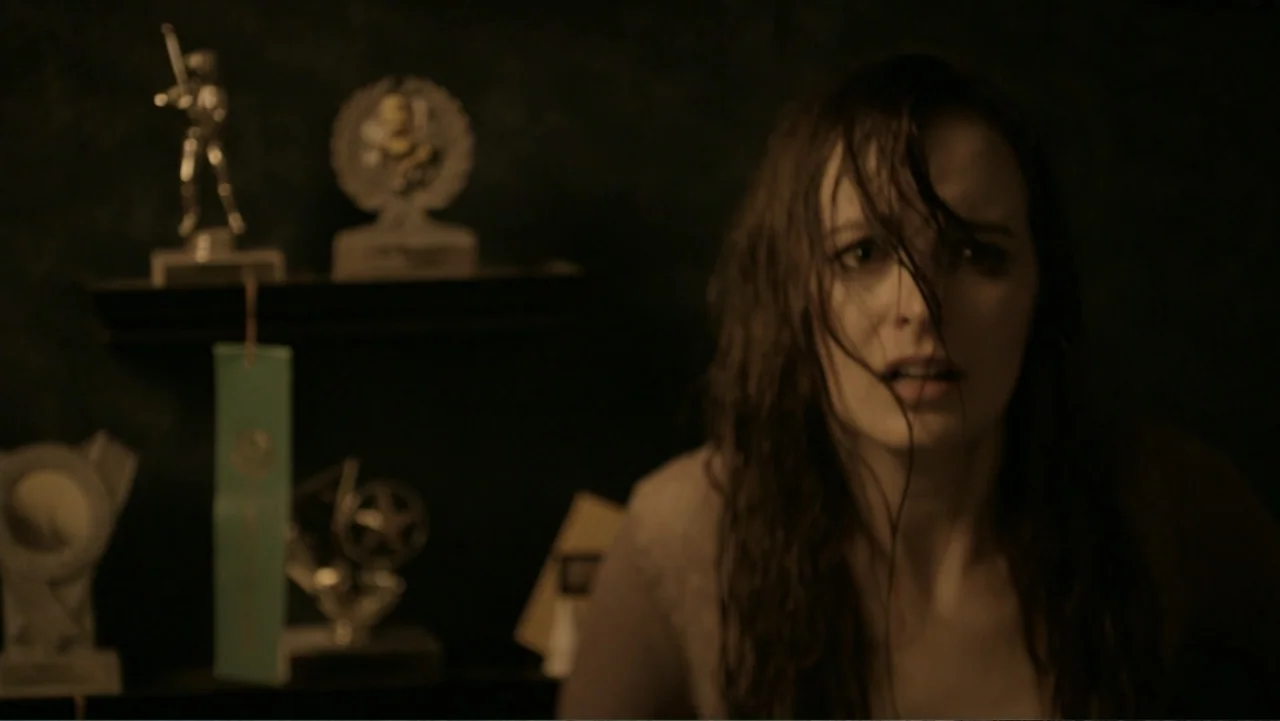 Totem (2022) movie still: closeup — A dimly lit room with several small trophies visible on a shelf in the background. A young woman with wet hair…