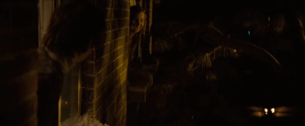 Let Me In (2010) movie still: medium — A brick house exterior at night shows a window with two people talking. One girl looks out from the frame…