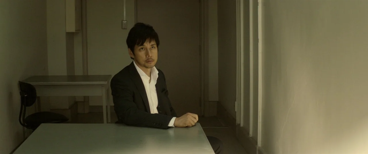 Creepy (2016) movie still: medium — An austere room features plain light-colored walls, a grey metal table, and a chair. A man in a dark suit sits…