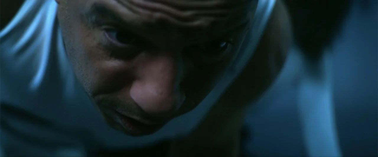 Bloodshot (2020) movie still: closeup — A dimly lit room with cool blue ambient light cast across the frame. A man in a white tank top leans forward…