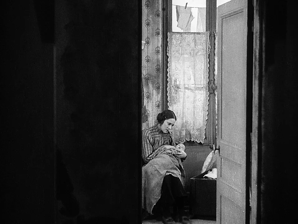 Coeur Fidele (1923) movie still: medium — A cluttered apartment room features patterned wallpaper and lace curtains over a window. A woman sits in a…