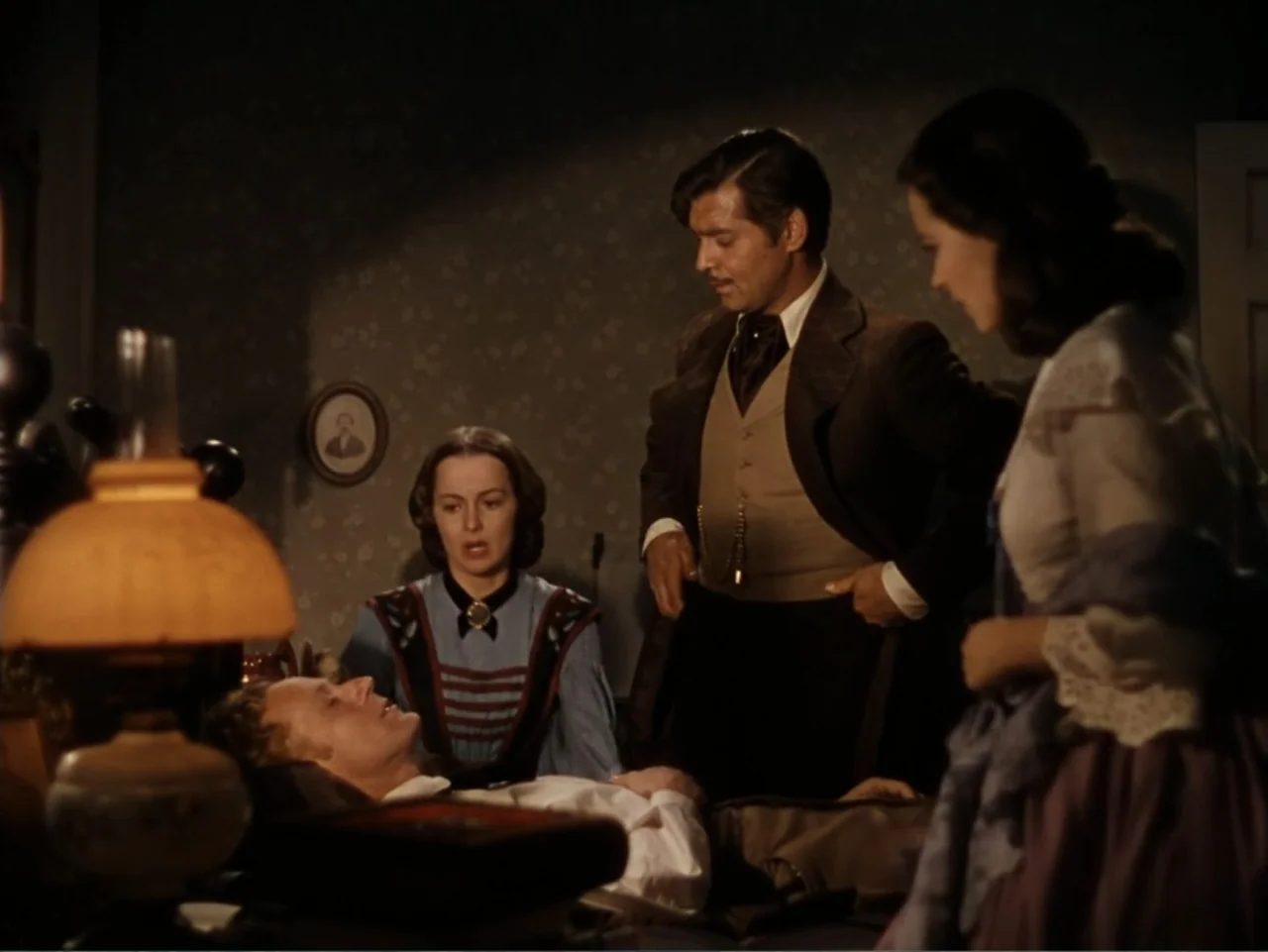 Gone with the Wind (1939) movie still: medium — An 1800s interior with floral wallpaper and a small portrait frame on the wall. A group stands over…