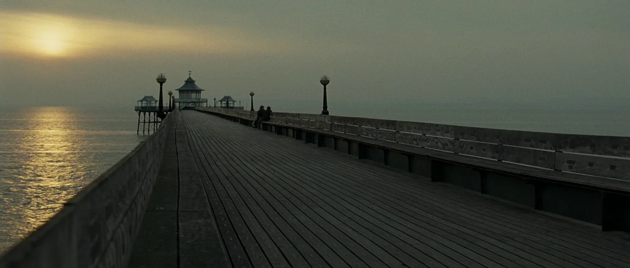 Never Let Me Go (2010) movie still: wide — A long wooden pier extends over calm water toward a structure in the distance. Two figures sit on a bench…