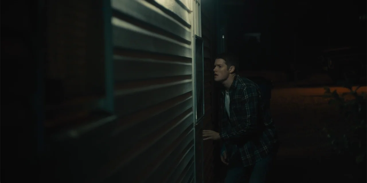 All Roads to Pearla (2019) movie still: medium — A exterior wall of a building features vinyl siding with a single light source. A young man stands…