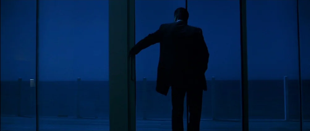 Heat (1995) movie still: A man in a suit stands silhouetted against a vast, deep blue expanse, peering through a modern glass door, possibly onto a…