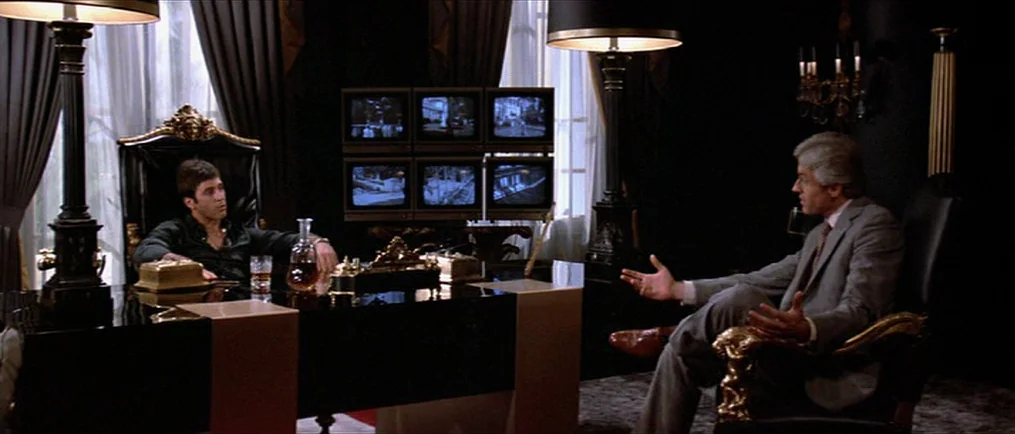 Scarface (1983) movie still: wide — A dark, luxurious home office features a large black desk and six security monitors. Two men sit in ornate…