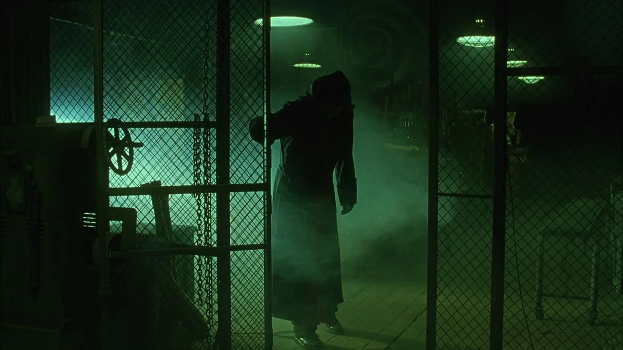 Saw movie still: fullbody — A dark, industrial storage area features metal mesh fencing and thick plumes of smoke. A single figure in a long dark…