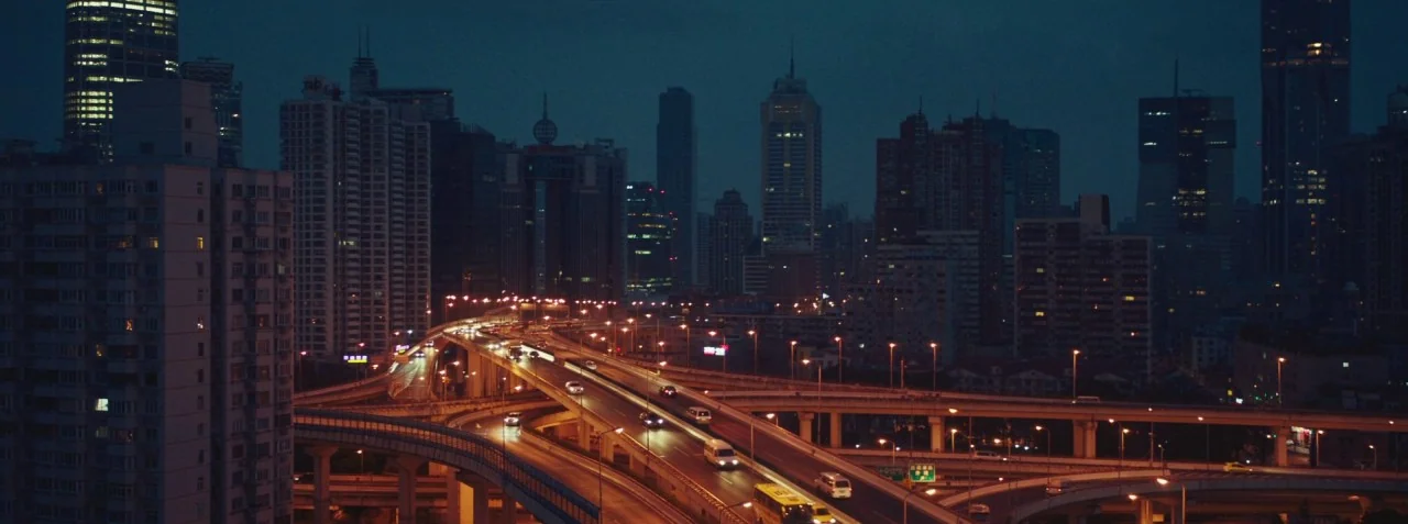 Dead Pigs (2018) movie still: establishing — A sprawling concrete highway interchange snakes through a dense urban skyline at night. Rows of tall…