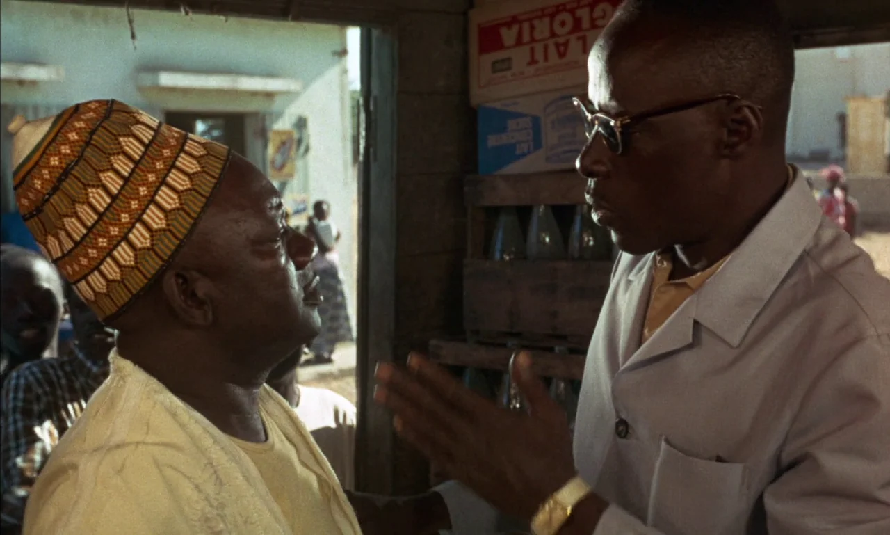 Mandabi (1968) movie still: medium — A small room containing stacked crates of glass bottles is lit by daylight. Two men stand facing each other…