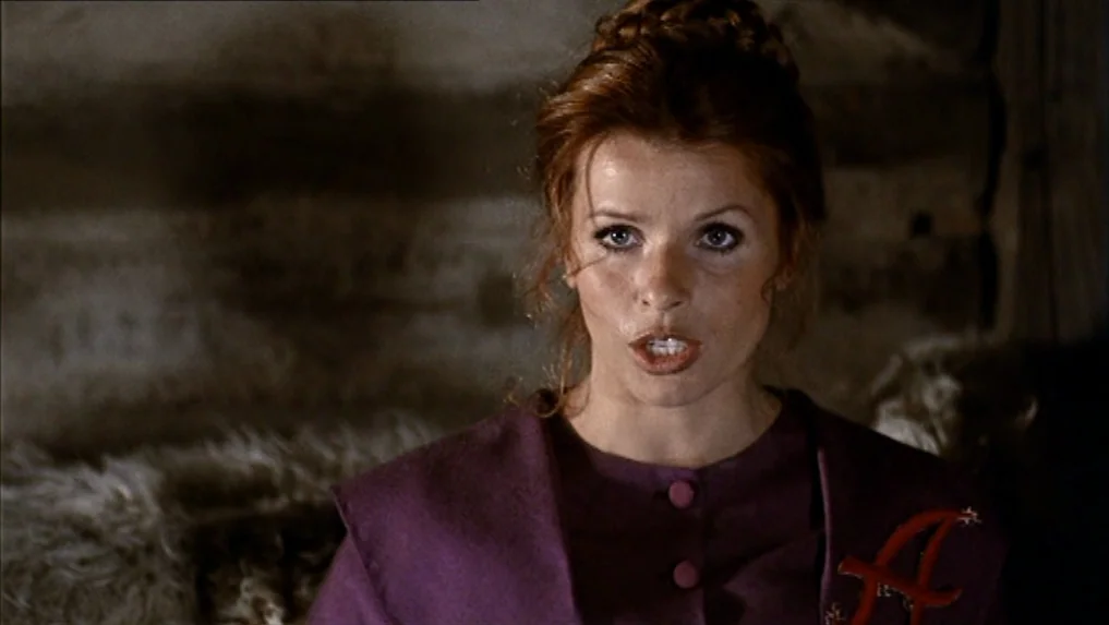 The Scarlet Letter (1973) movie still: closeup — A dimly lit wooden interior with rough-hewn log walls and dark fur textures. A young woman with…