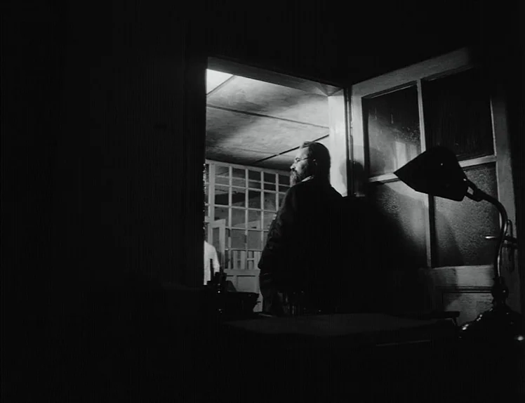 Damnation (1988) movie still: medium — A dimly lit room with a dark foreground features an open rectangular doorway. A bearded man in a suit stands…
