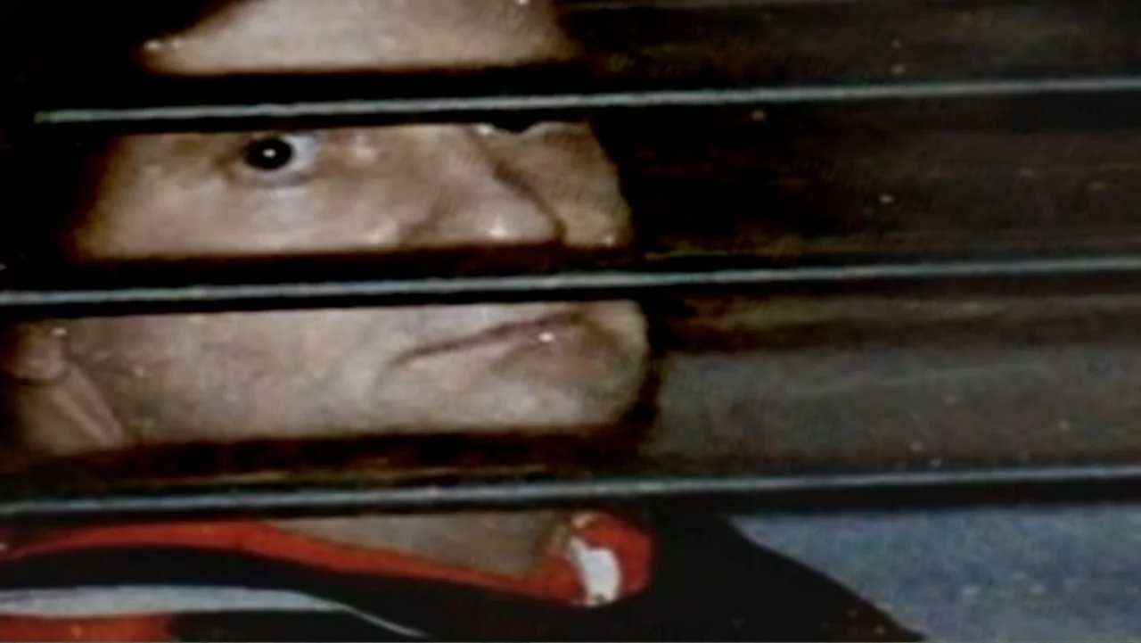 The Cheshire Murders (2013) movie still: closeup — A man's face is partially obscured by horizontal venetian blinds in a dimly lit room. The man…