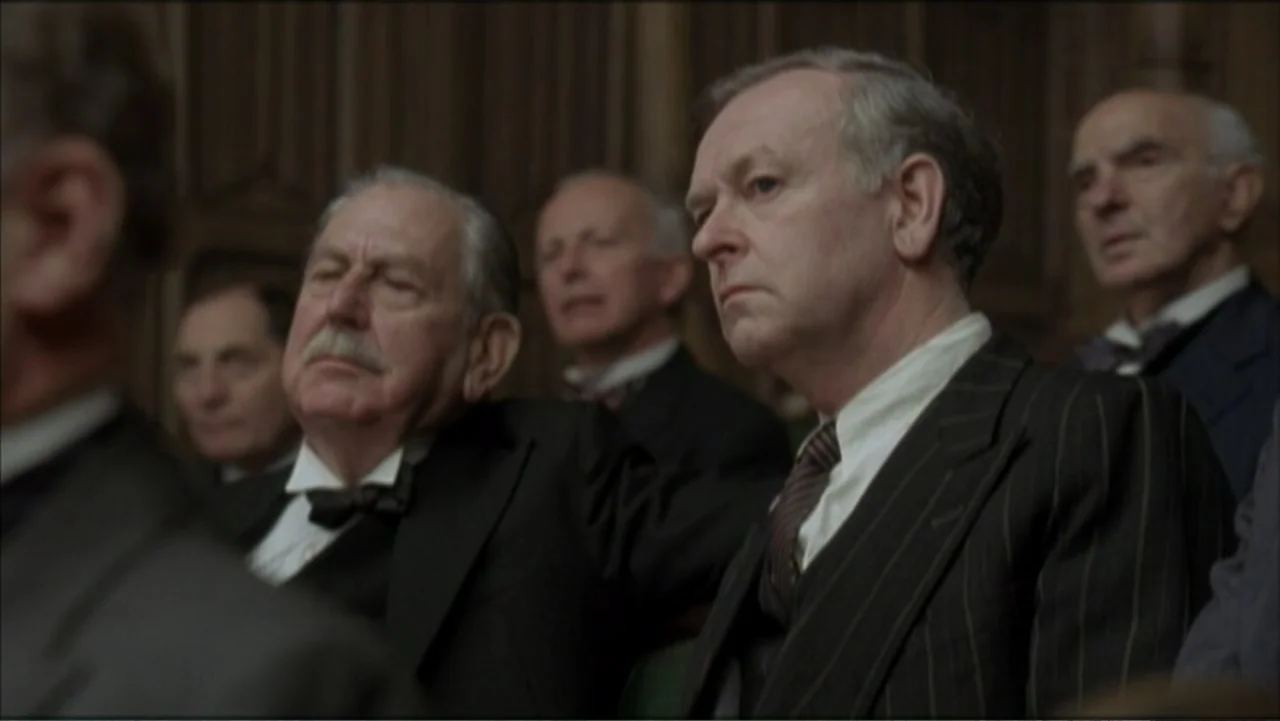 The Gathering Storm (2002) movie still: medium — An interior space features wood paneled walls behind several men seated in rows. The men wear…