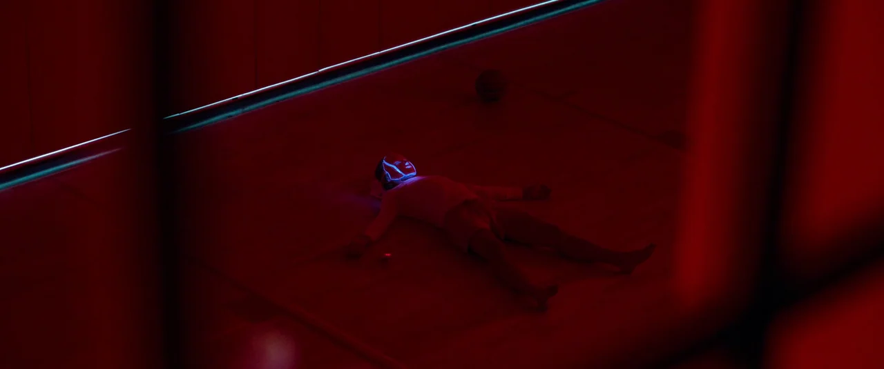 Bodies Bodies Bodies (2022) movie still: wide — A dimly lit indoor basketball court is bathed in deep red light. A lone person wearing a glowing…