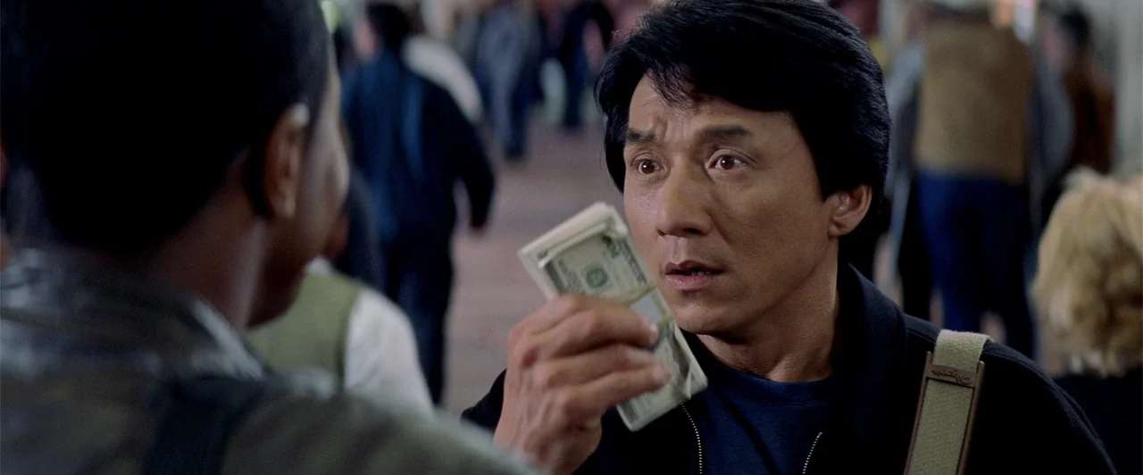 Rush Hour 2 (2001) movie still: medium — A crowded public outdoor area with blurry figures in the background. A man holds a thick bundle of cash…