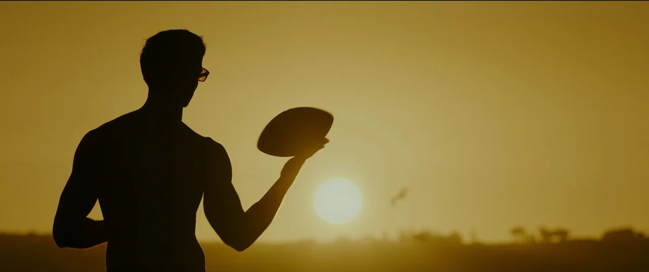 Top Gun: Maverick (2022) movie still: medium — A silhouette of a man stands on a beach during a vibrant sunset. He holds a football out in front of…