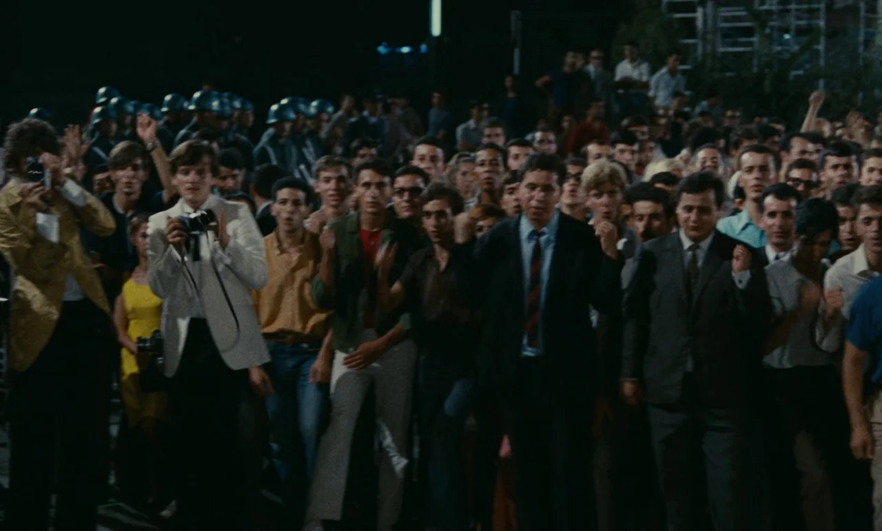 Z (1969) movie still: medium — A dense crowd of people walks forward in a dark, dimly lit urban street. Several individuals carry cameras while…